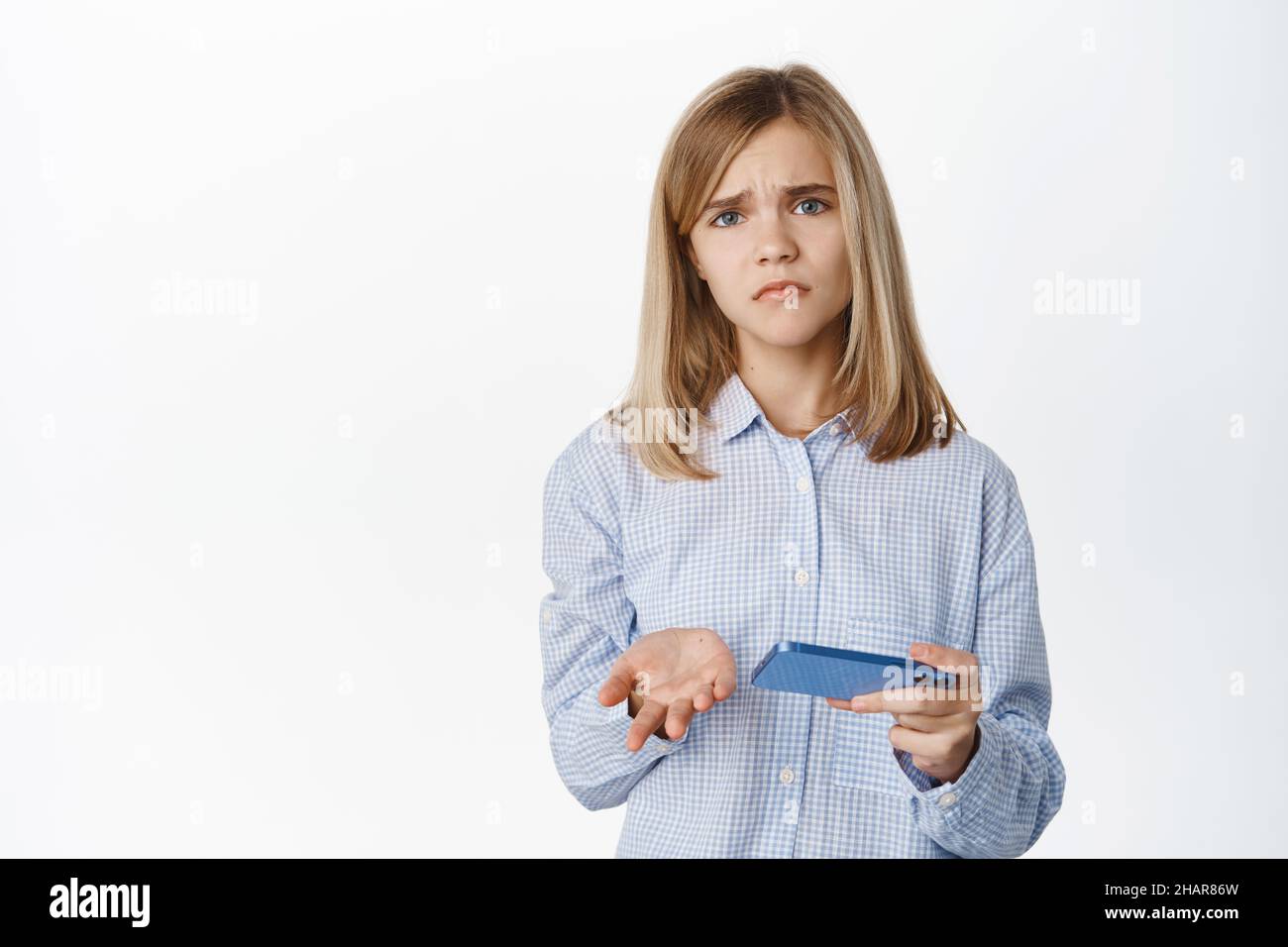 Sad and disappointed girl holding smartphone and looking upset, losing in video game, watching video on mobile phone, standing over white background Stock Photo