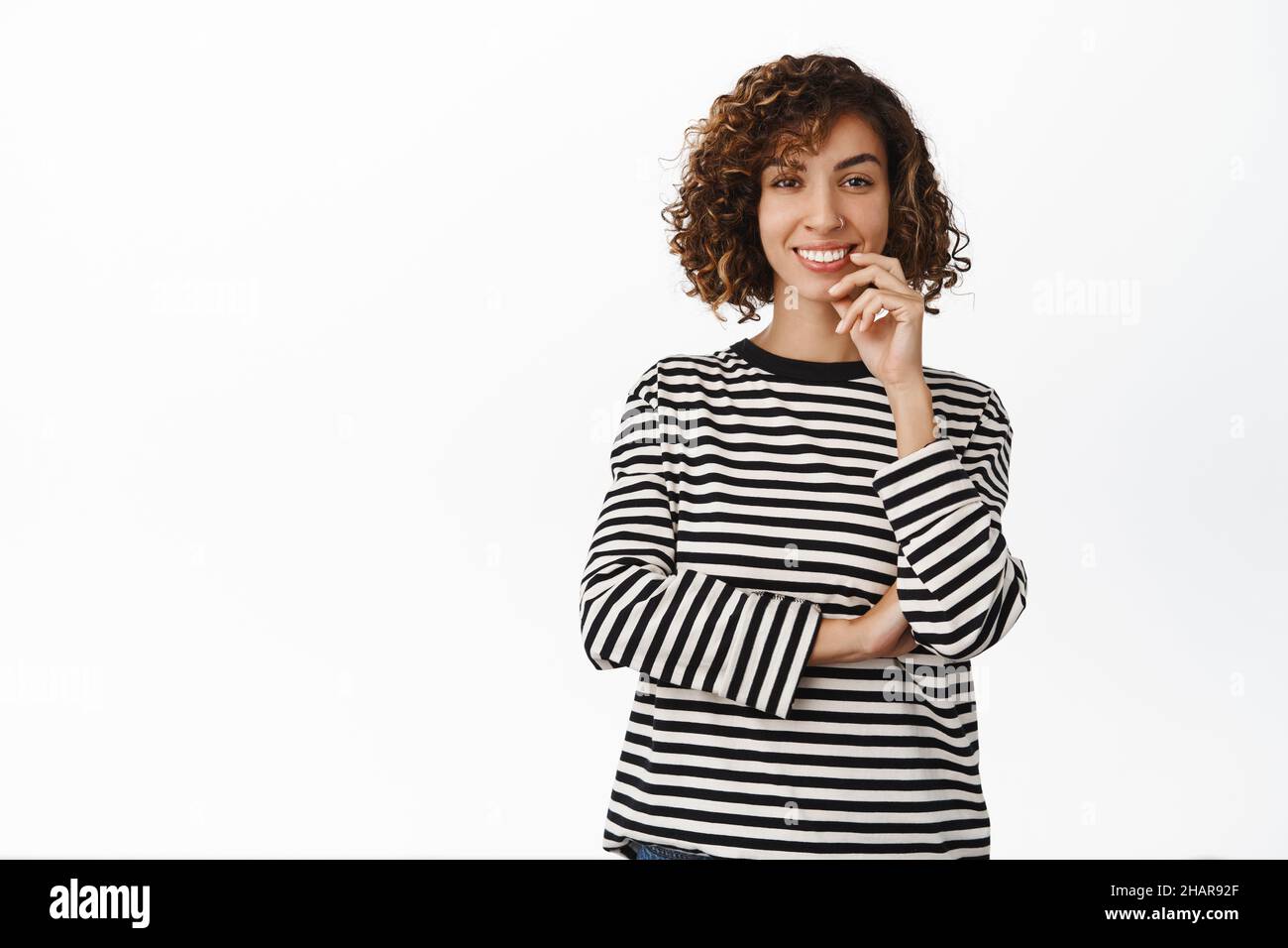 Portrait of stylish, attractive young middle eastern woman with curly short hair, smiling at camera and looking thoughtful, white background Stock Photo