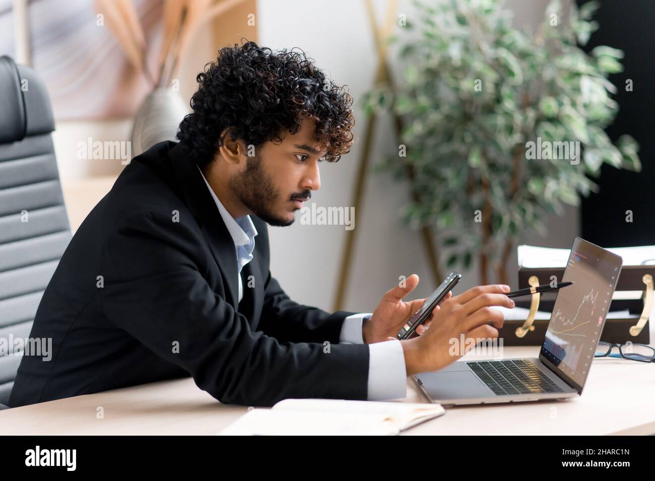 Successful confident focused purposeful Indian male trader, uses mobile phone and laptop to buy and sell cryptocurrency, analyzes financial market of cryptocurrencies, invests in electronic currency Stock Photo
