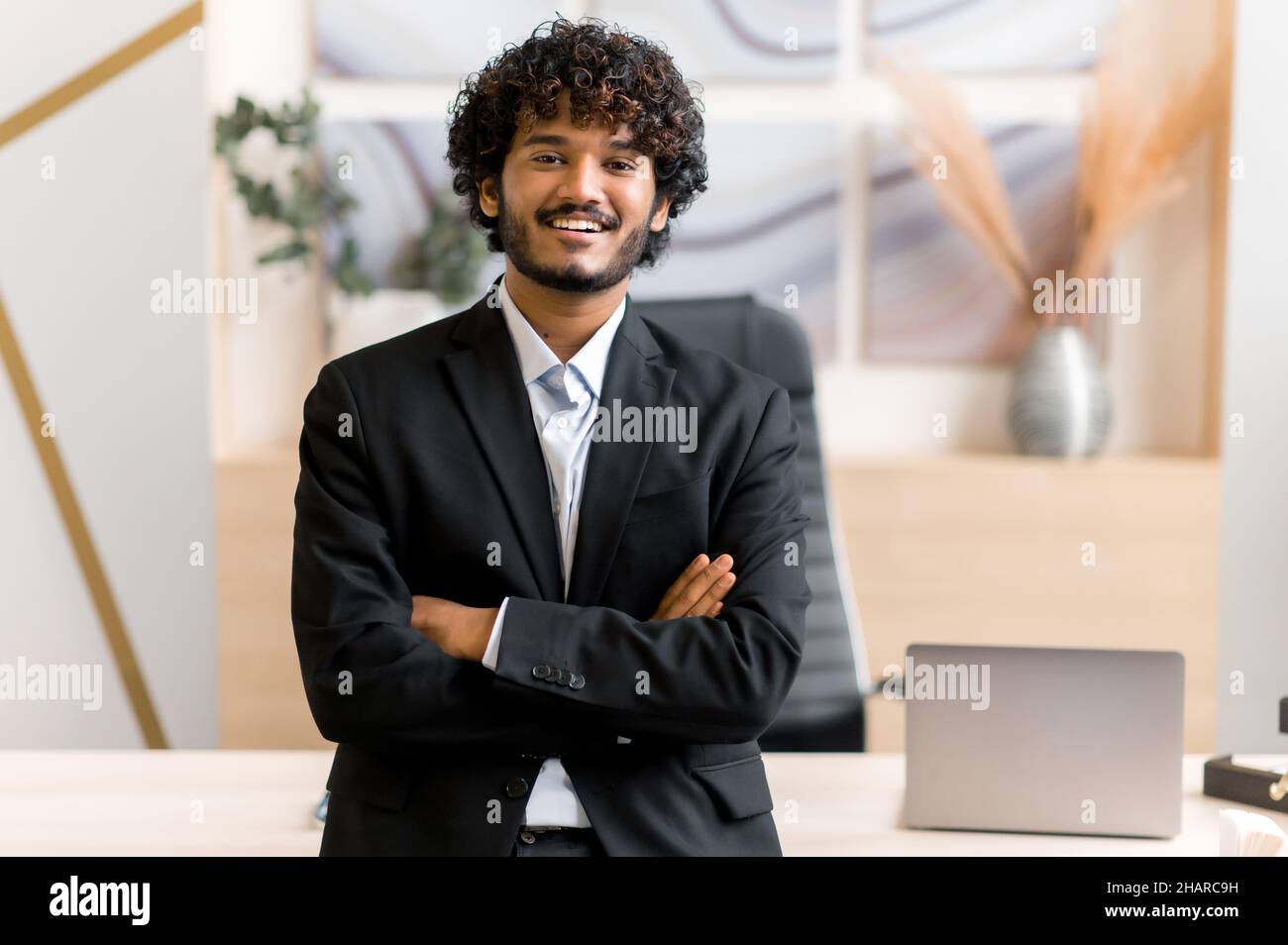 Portrait successful attractive stylish young adult Indian business leader, manager or IT professional dressed in formal suit standing near his desk in office with arms crossed, look at camera, smile Stock Photo