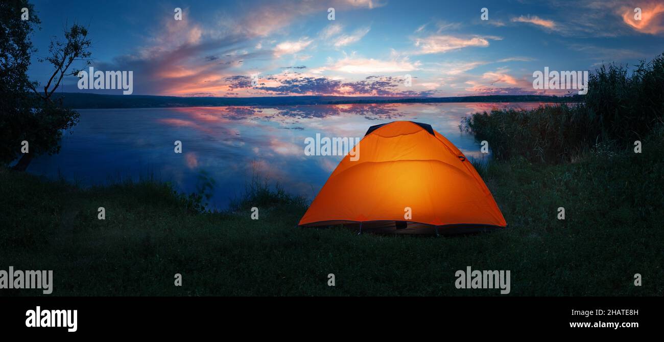 Internally lit orange tent on shore of lake under dramatic sunset sky Stock Photo