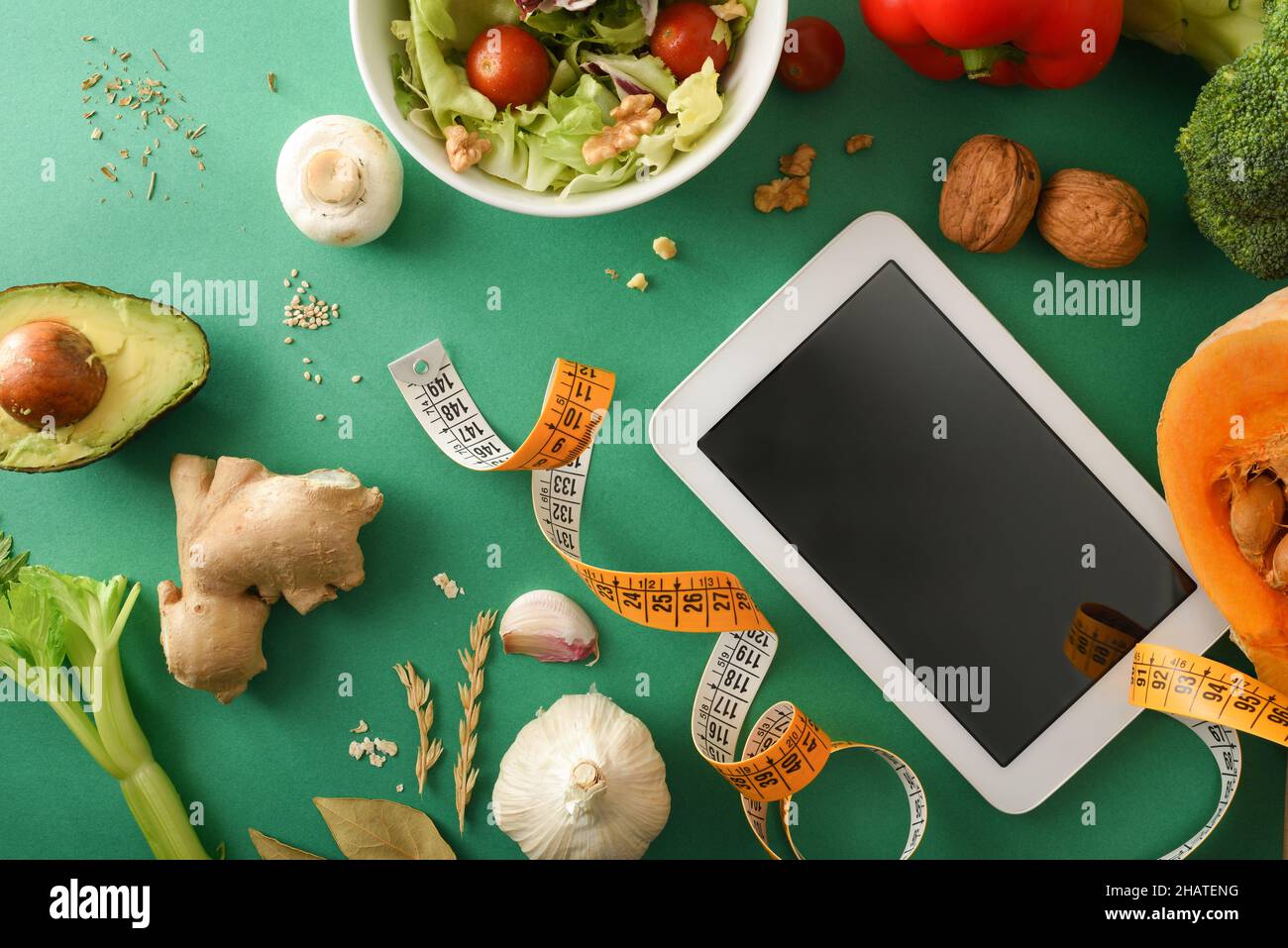 Weight control planning with blank electronic device on green table for preparation of healthy dishes with vegetables and measuring tape around. Top v Stock Photo