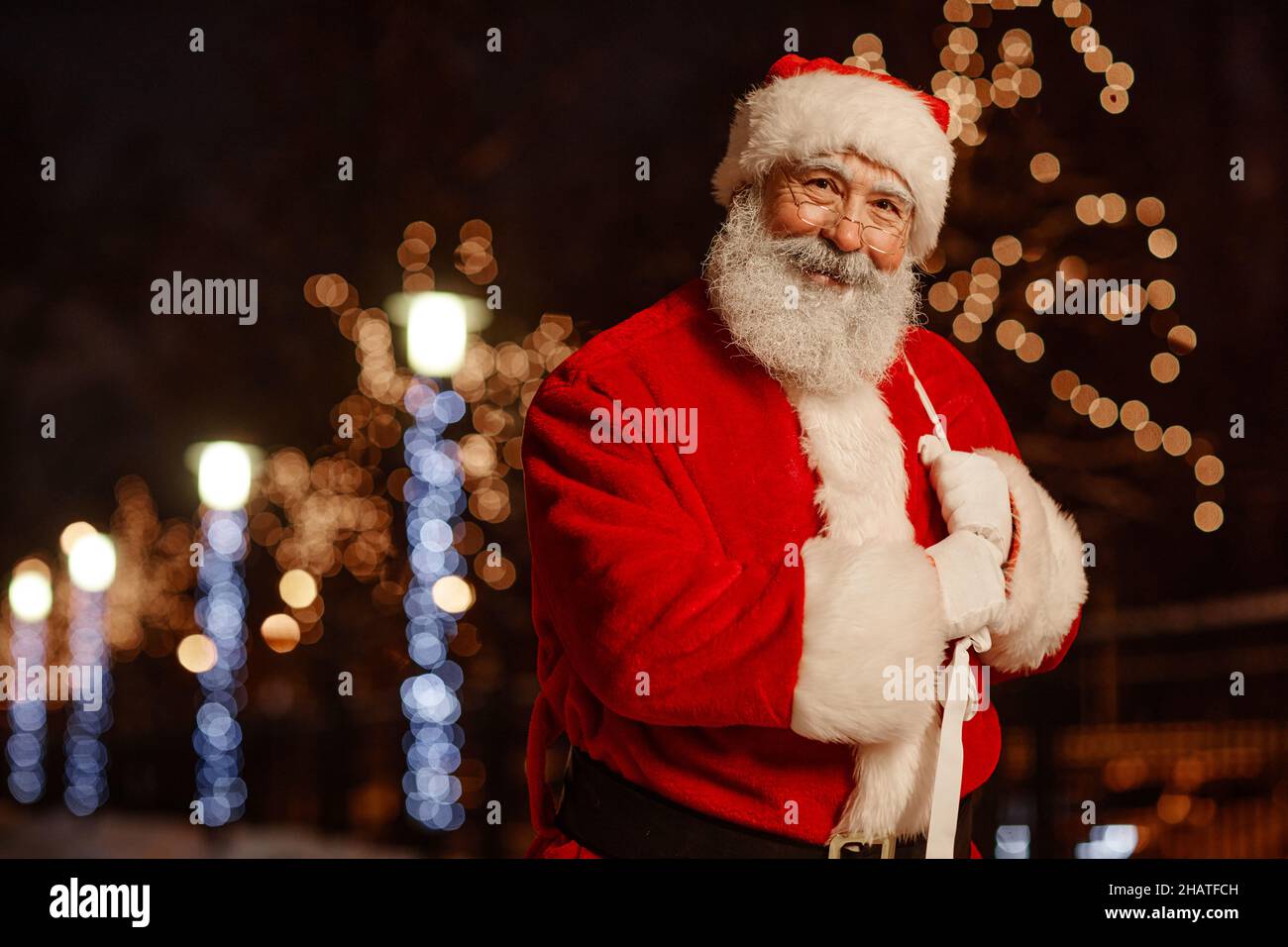 Horizontal medium portrait of joyful Santa Claus standing outdoors in street decorated with garland lights smiling at camera, copy space Stock Photo