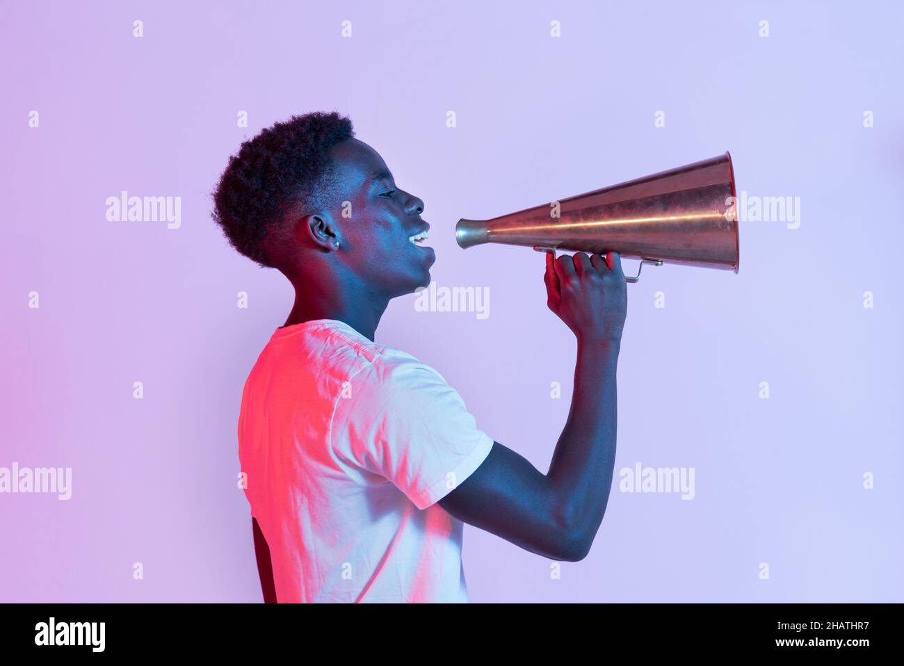 Side view of cheerful young black male millennial in t shirt smiling while talking with metal vintage megaphone against purple background Stock Photo