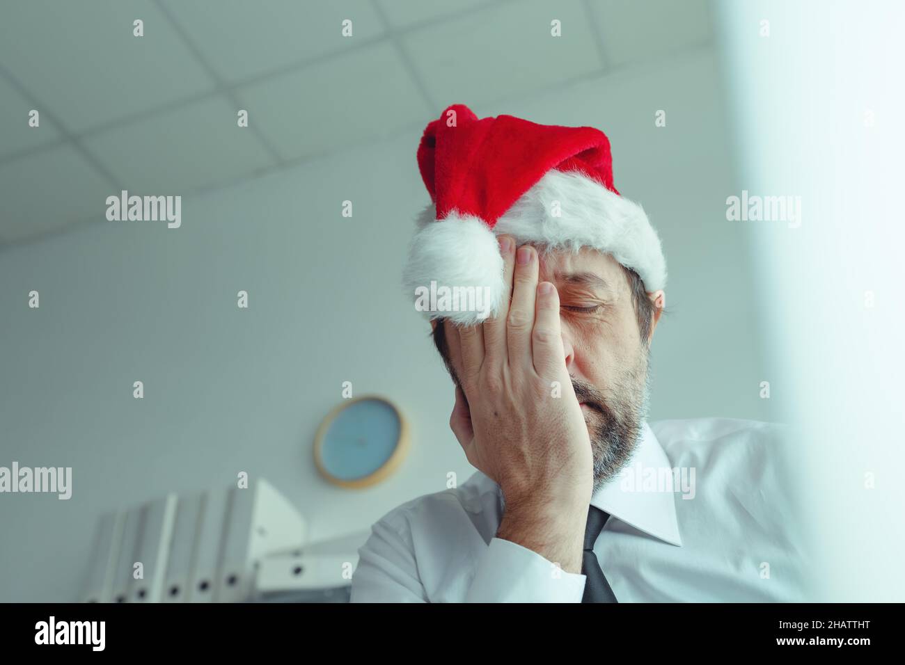 Pessimistic businessman with Santa Claus hat suffering from Christmas holiday season depression in office, selective focus Stock Photo