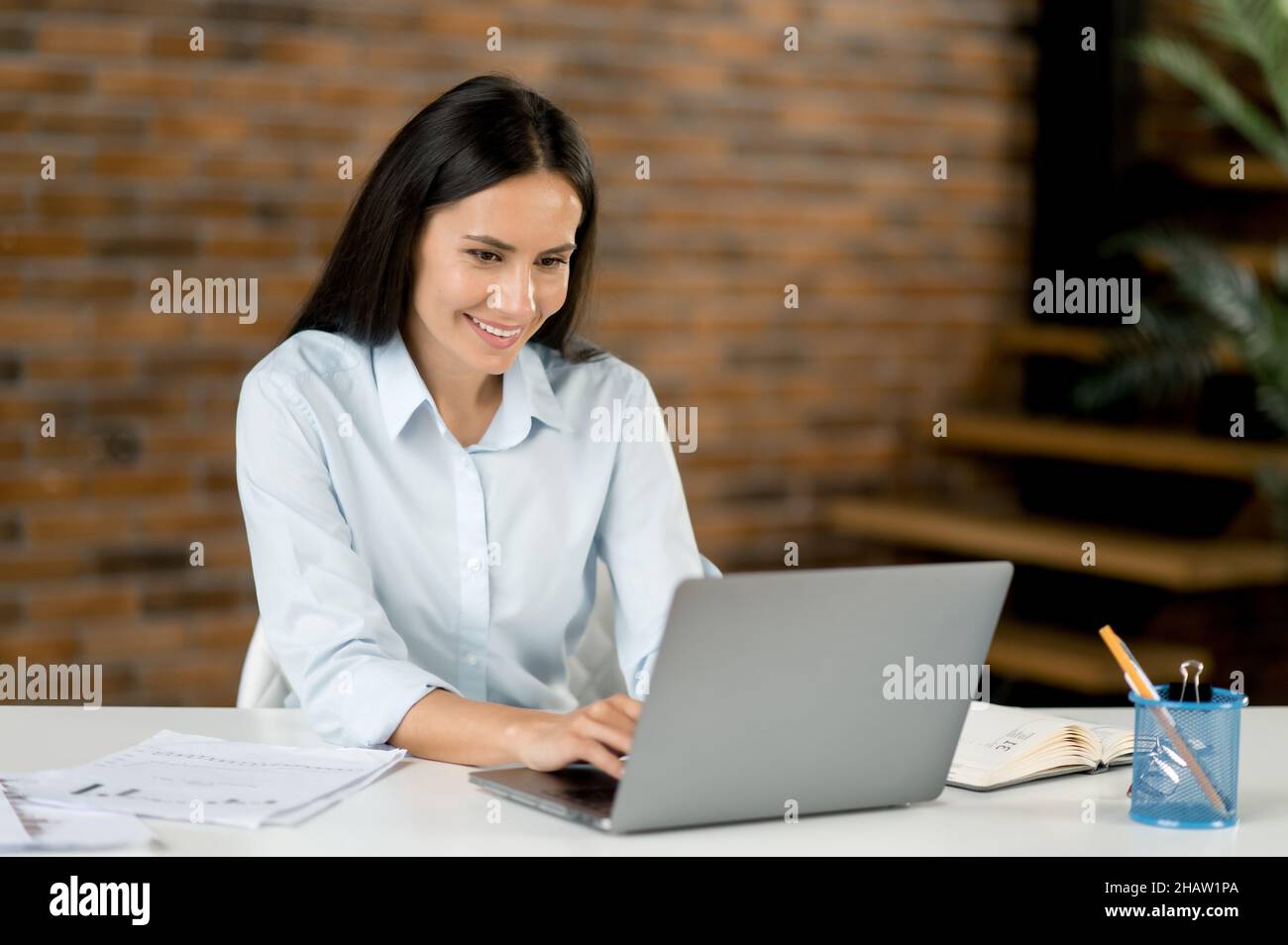 Portrait caucasian young business lady in office. Attractive confident smart woman top manager or trader, brunette, uses laptop for text messages with colleagues or client, prepares business project Stock Photo
