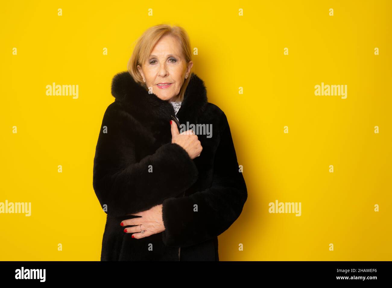 Portrait of elderly woman wearing winter clothing feeling cold on yellow background Stock Photo