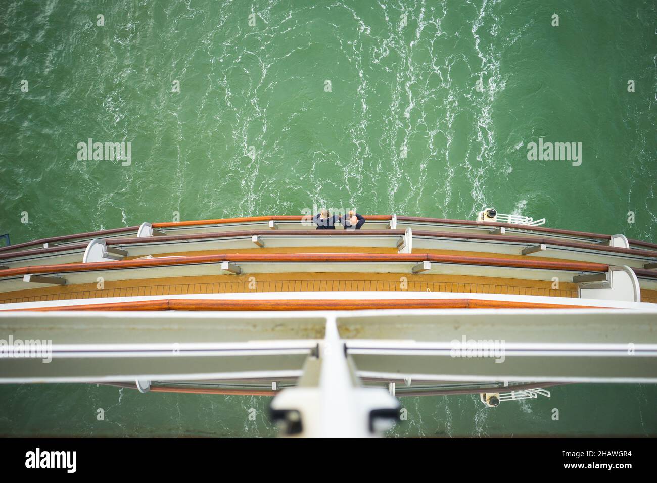 Looking down at a couple standing on the balcony at the rear of a cruise ship, showing the stacked rails of the decks and the green sea below Stock Photo