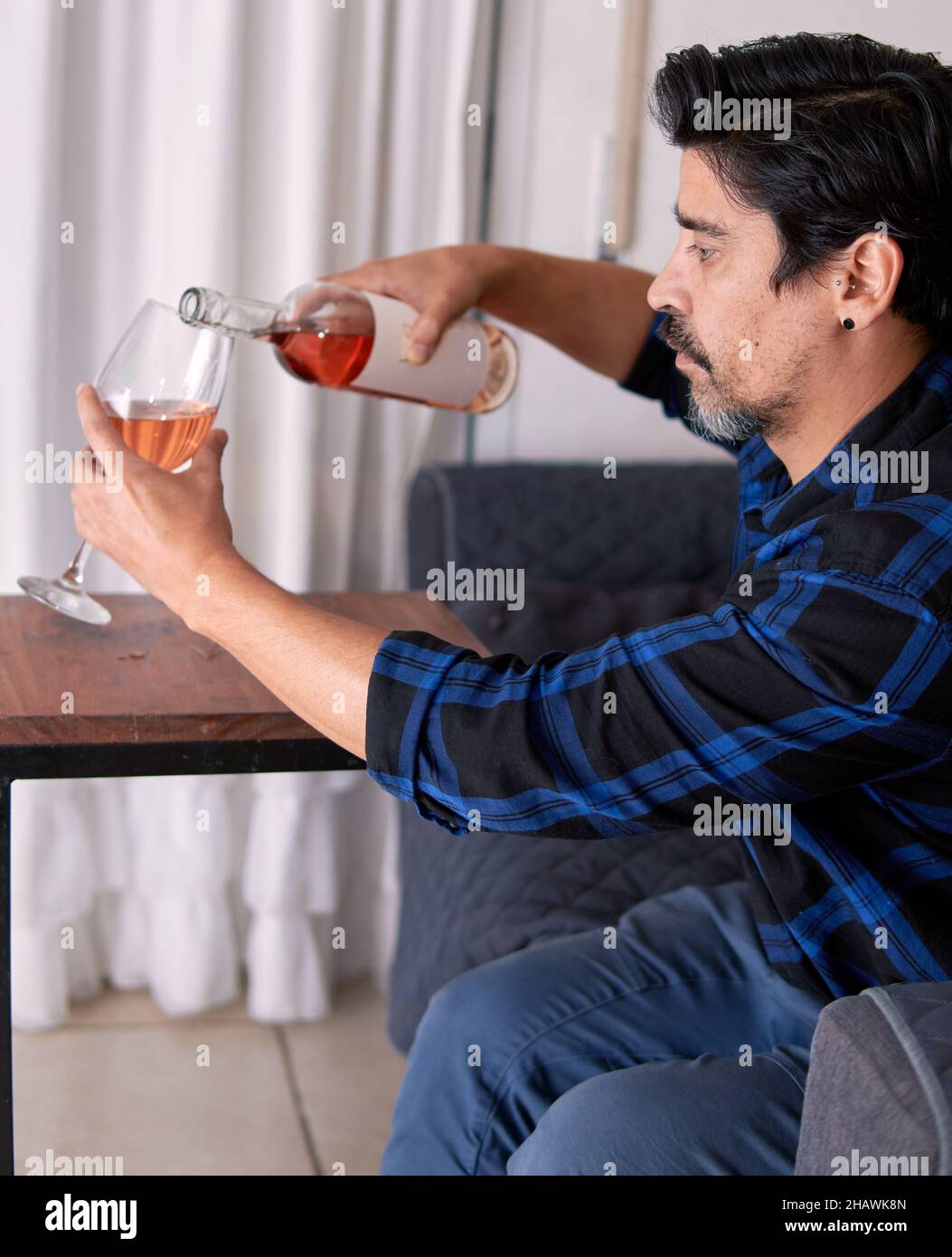 brunette latin man with shirt and beard pouring rose wine into a glass on a Table Wheel Sofa Side. White Curtain in background. Argentina. Vertical Stock Photo