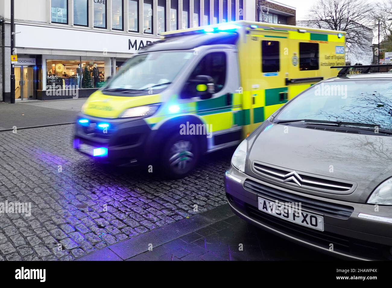 Motion Blur front & side view East of England NHS ambulance on blue lights emergency call driving at speed wet cobbled road Brentwood High Street UK Stock Photo