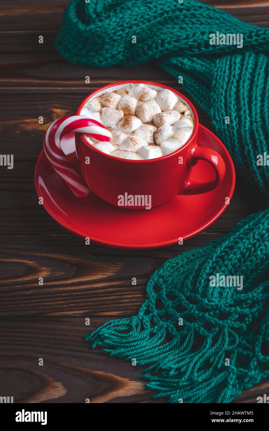 Red cup of cocoa with marshmallows and candy cane. Hot chocolate and warm knitted scarf on a dark wooden table. Winter season, card, minimal design. C Stock Photo