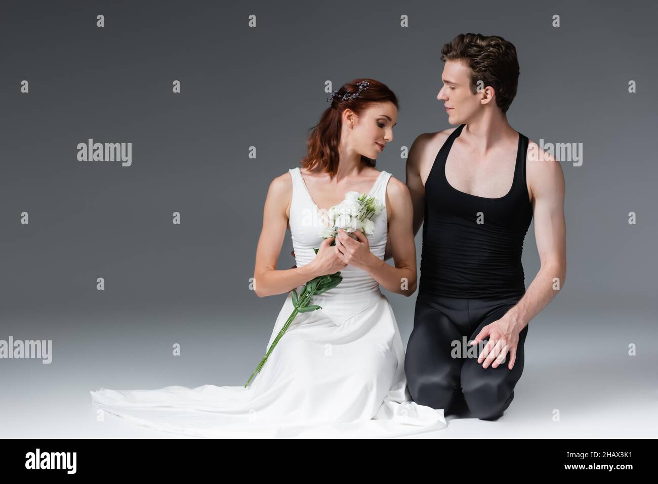 graceful ballerina in white dress holding flowers and standing on knees with partner on dark grey Stock Photo