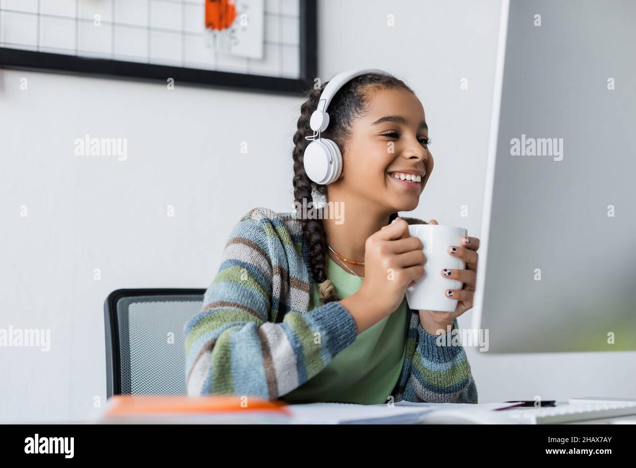 happy african american schoolgirl in headphones holding tea cup while looking at monitor Stock Photo