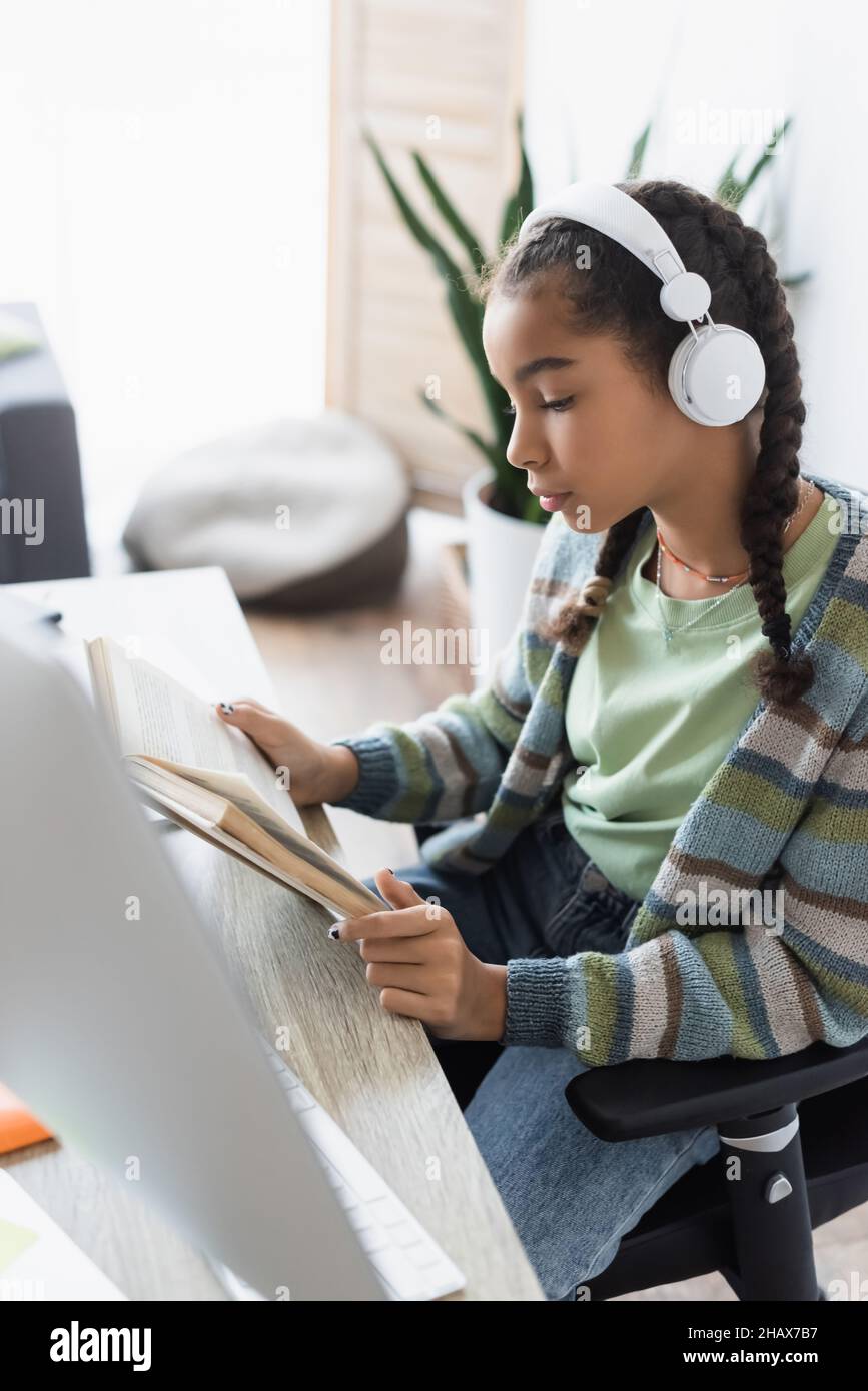 african american schoolgirl in headphones reading book near blurred computer monitor Stock Photo