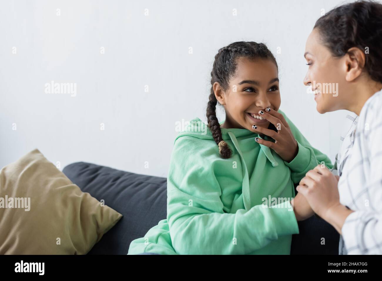 laughing african american girl covering mouth with hand while talking with mom at home Stock Photo