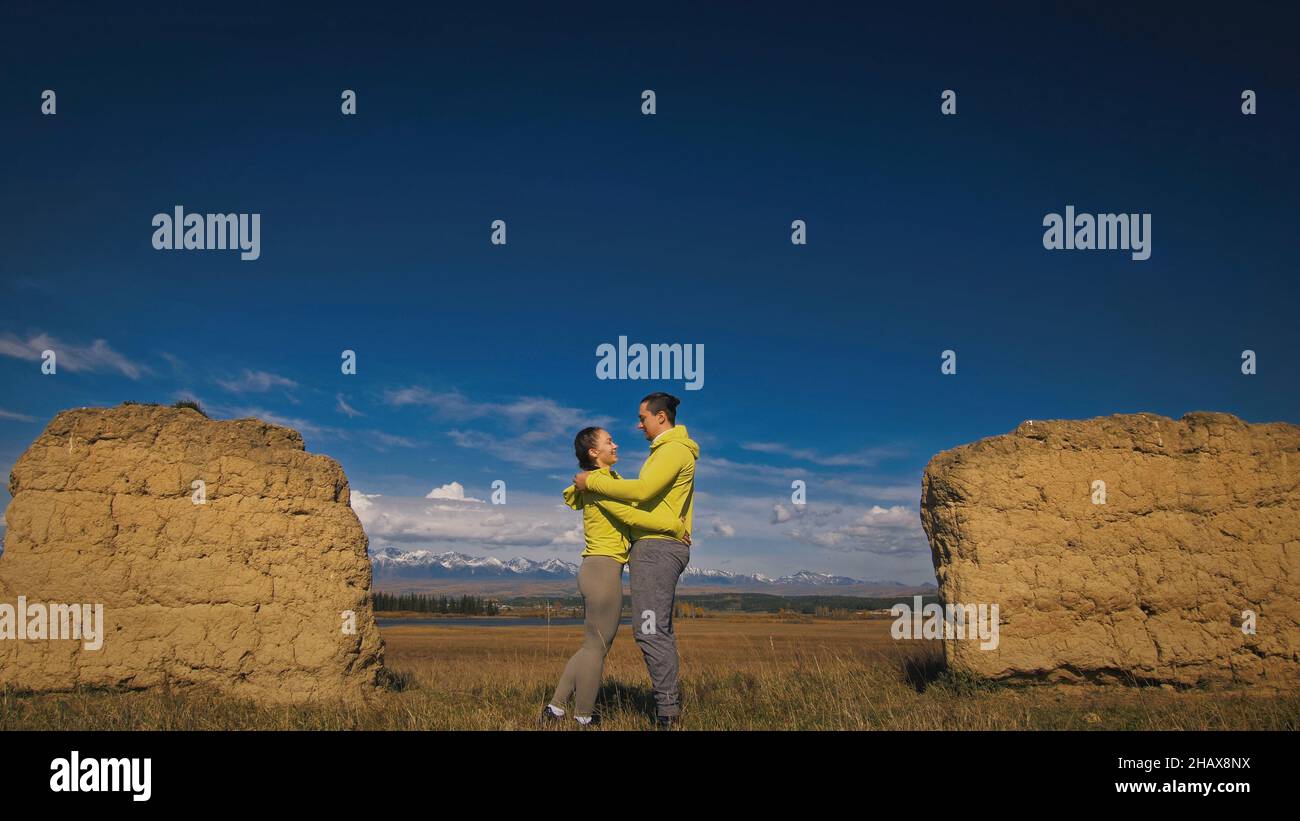 Man and woman in yellow green sportswear. Lovely couple of travelers hug and kiss near old stone enjoying highland landscape. Two travelers are walkin Stock Photo