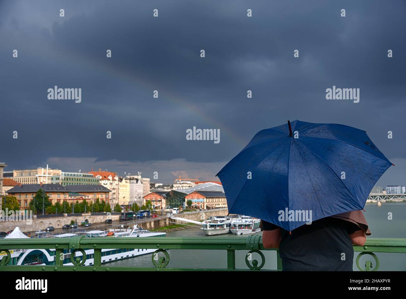 Person standing on a bridge under umbrella watches rainbow over the city. Rear view Stock Photo