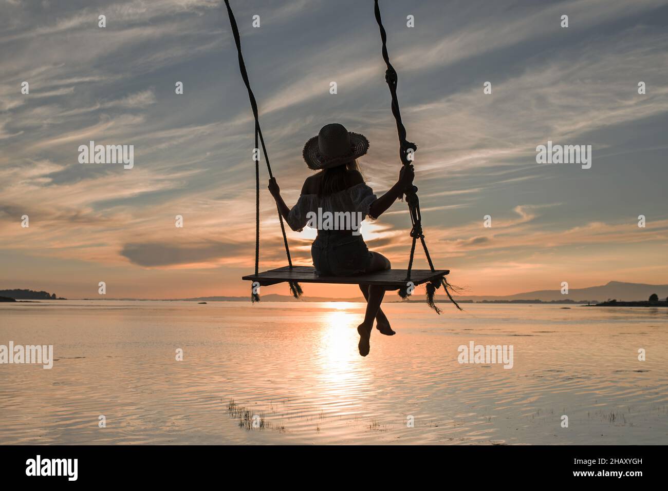 Back view of silhouette of anonymous young female in hat sitting on swing and enjoying amazing sundown sky in Cambados in Spain Stock Photo