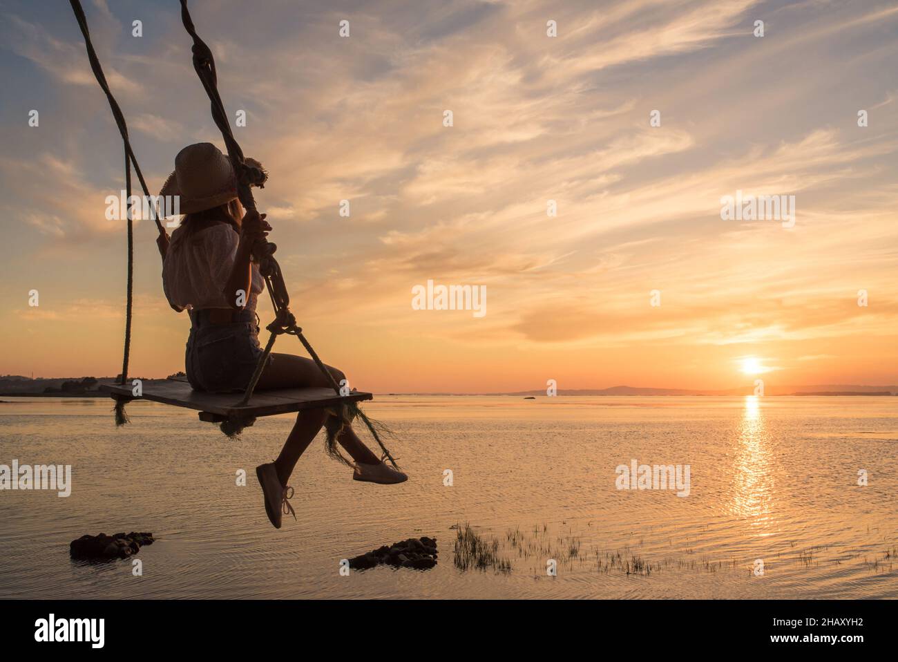 Back view of silhouette of anonymous young female in hat sitting on swing and enjoying amazing sundown sky in Cambados in Spain Stock Photo