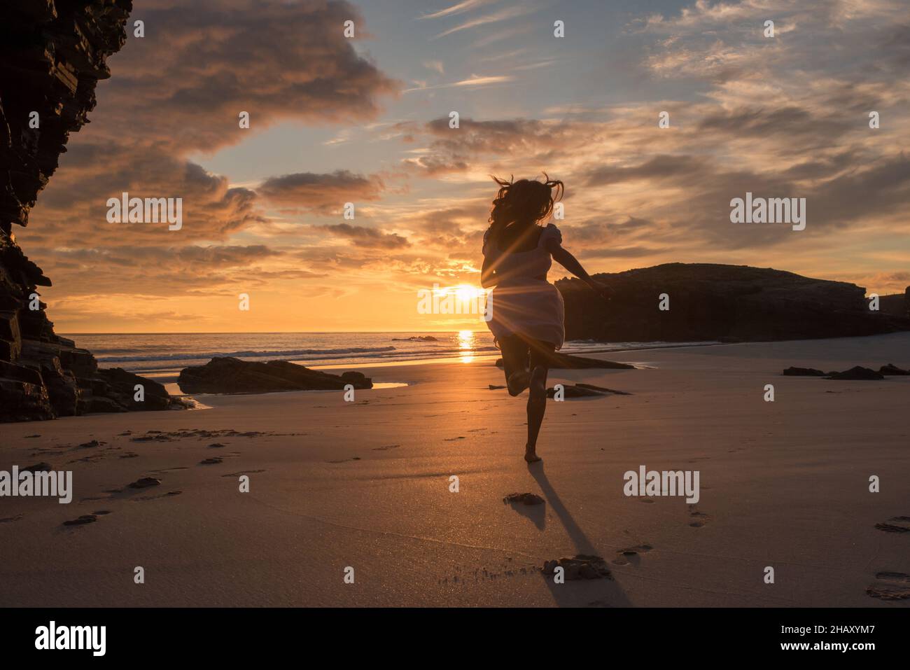 Silhouette of anonymous female in dress jogging on sandy beach of Cathedrals near calm sea and enjoying sunset cloudy sky Stock Photo
