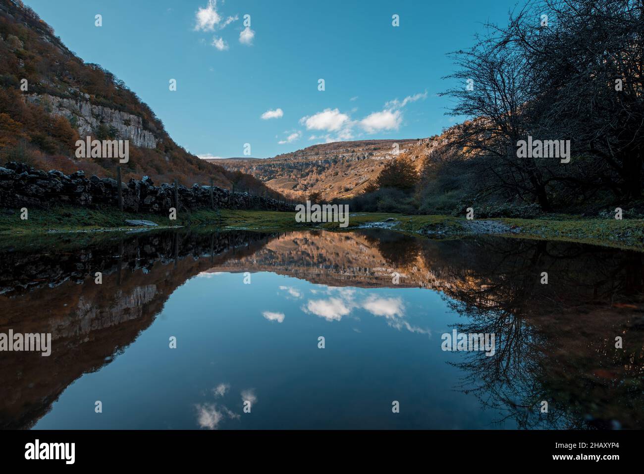 Picturesque view of calm lake surrounded by trees and and grass against Cantabria mountains in autumn under blue sky Stock Photo