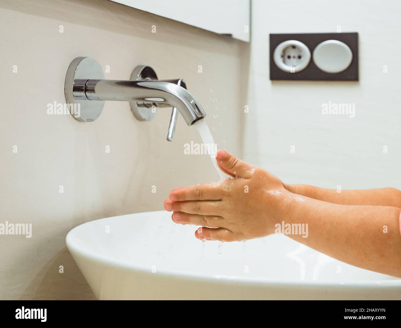 Crop anonymous kid washing hands at sink with pouring water from tap during daily hygiene routine in light bathroom at home Stock Photo