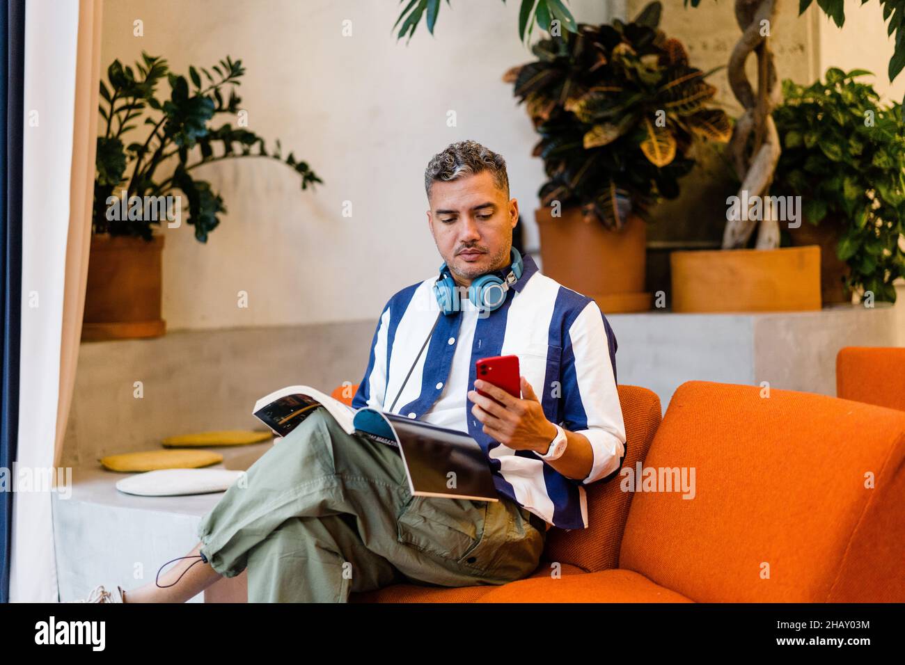 Focused male worker with headphones reading magazine and browsing on smartphone while sitting on armchair in modern office during work Stock Photo