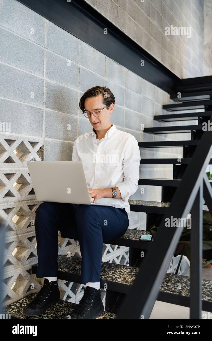 Focused male worker with netbook sitting on staircase while working in light modern workspace during work Stock Photo