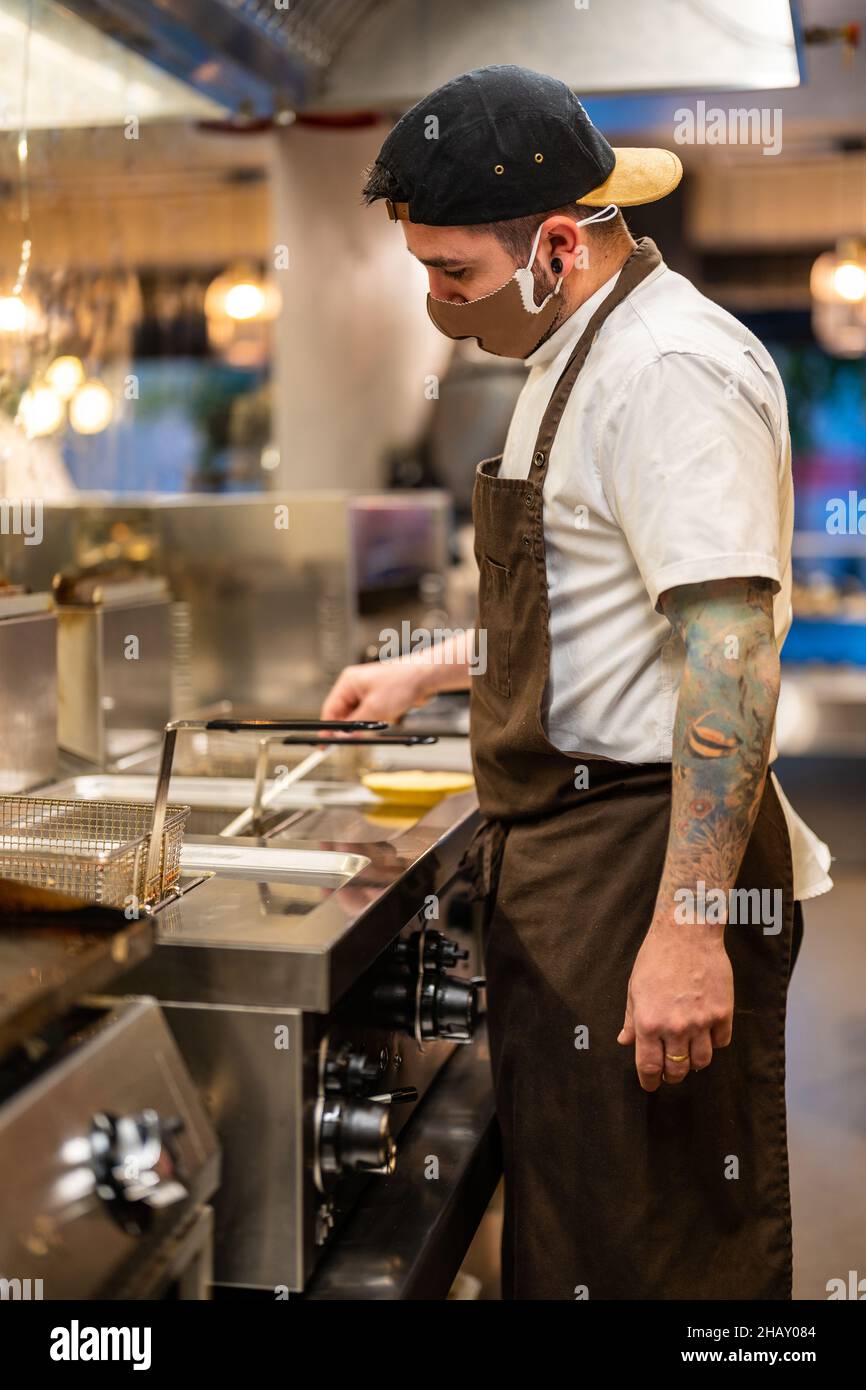 Side view of tattooed cook in uniform preparing food in deep fryer with boiling oil in kitchen of restaurant during coronavirus pandemic Stock Photo
