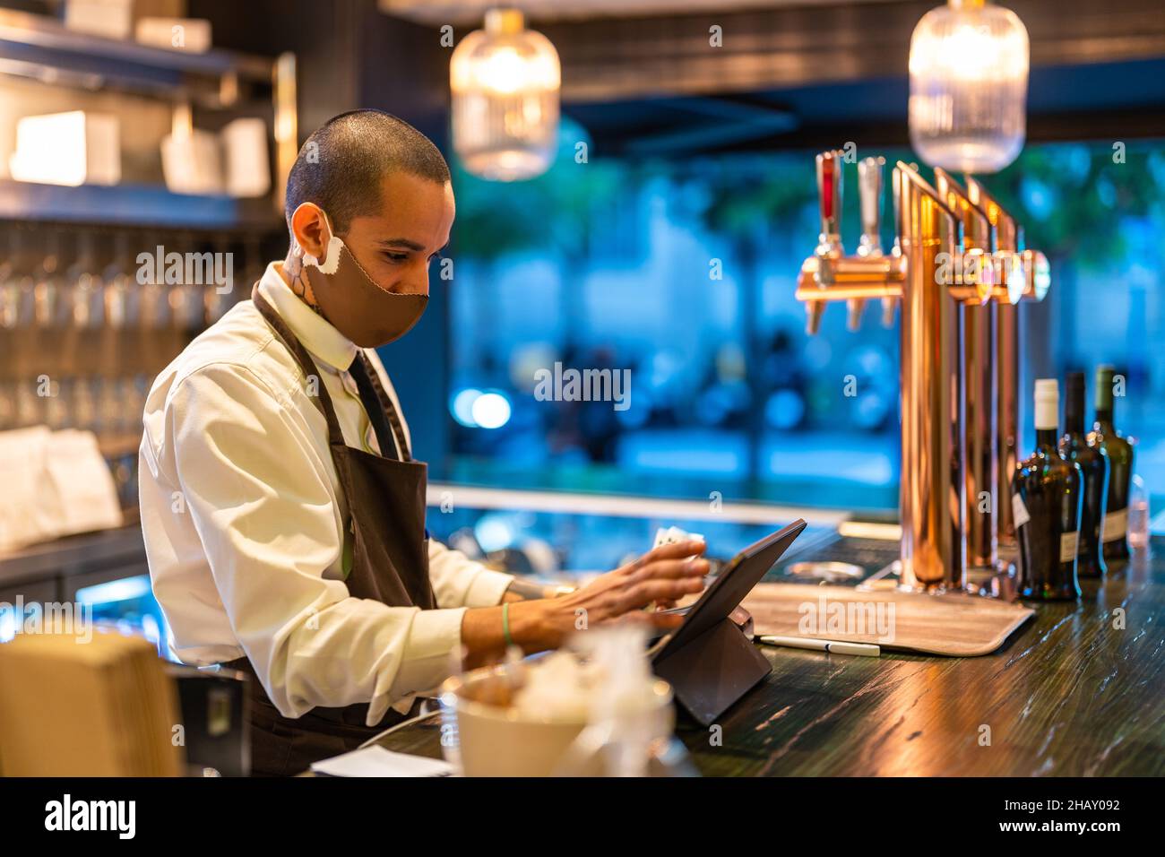 Serious male barkeeper in uniform and mask working on tablet at counter of bar during coronavirus pandemic Stock Photo