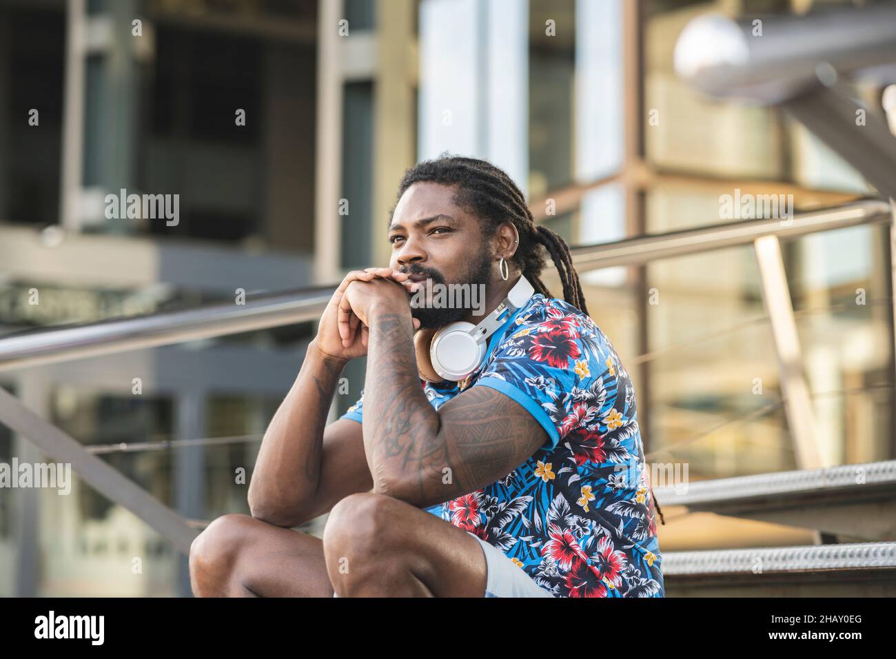 Thoughtful African American male in casual clothes with wireless headphones on neck looking into distance while sitting on stairs near building Stock Photo