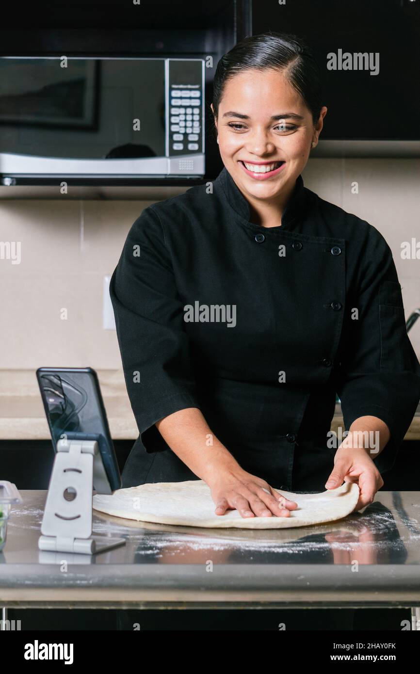Young smiling ethnic female chef in uniform taking video on smartphone while cooking delicious pizza on kitchen counter Stock Photo