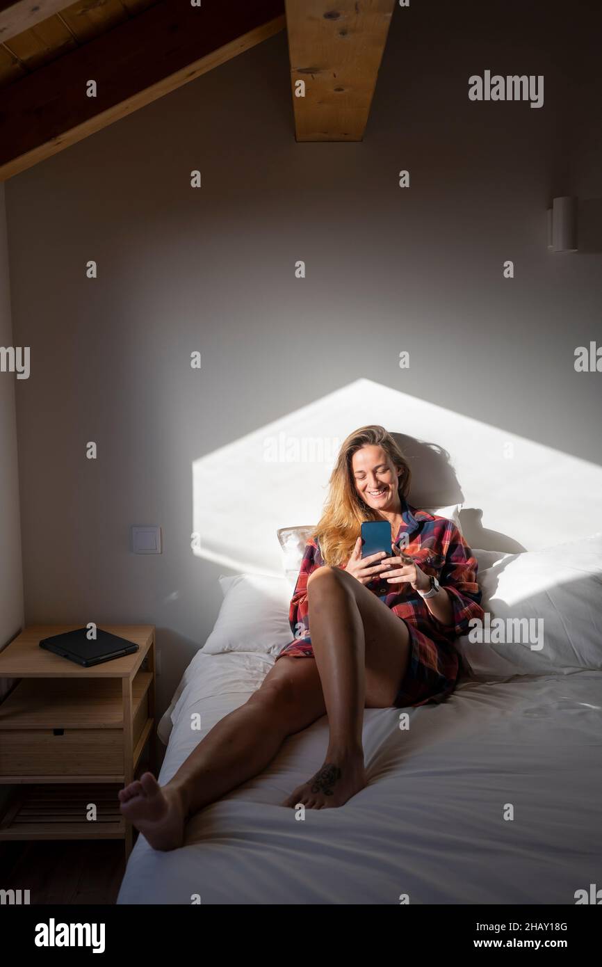 Blond lady sitting on bed and browsing smartphone while resting in attic Stock Photo