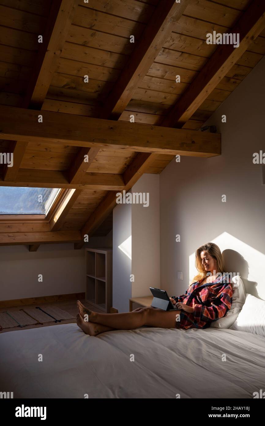 Side view of blond lady sitting on bed and browsing on tablet while resting in attic Stock Photo