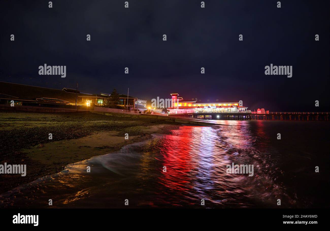Felixstowe, Suffolk, UK: Felixstowe pier at night illuminated with red and yellow lights. View of the pier from the beach with reflections in the sea. Stock Photo