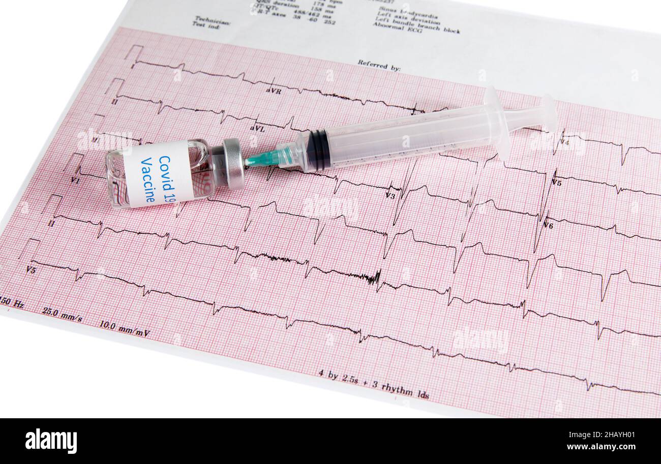 A Covid-19 vaccine bottle with a hypodermic needle inserted into the bottle with a syringe laying on top of a heart monitor graph or echocardiograph p Stock Photo