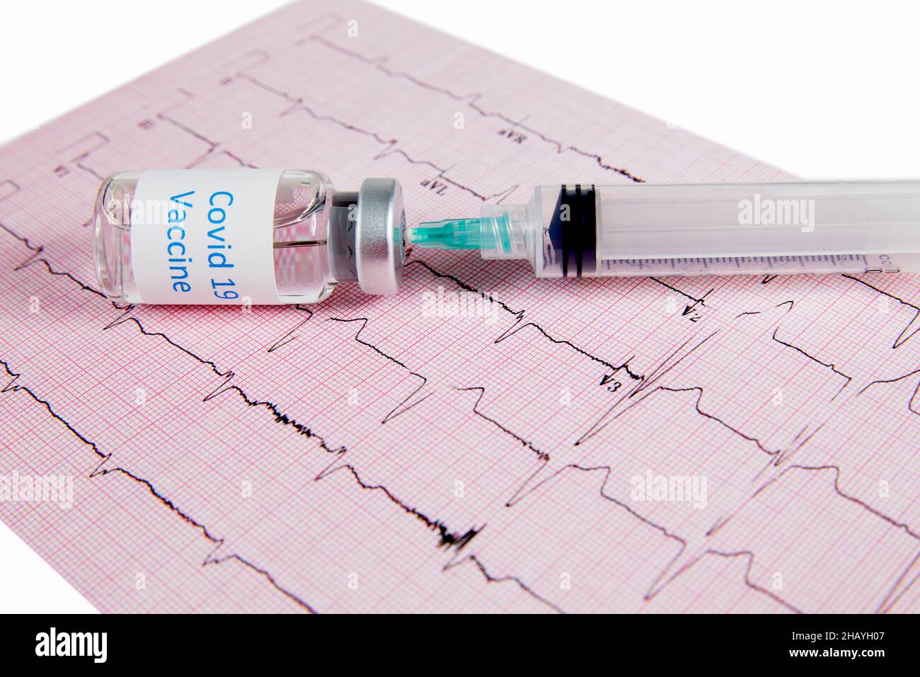 A Covid-19 vaccine bottle with a hypodermic needle inserted into the bottle with a syringe laying on top of a heart monitor graph or echocardiograph p Stock Photo