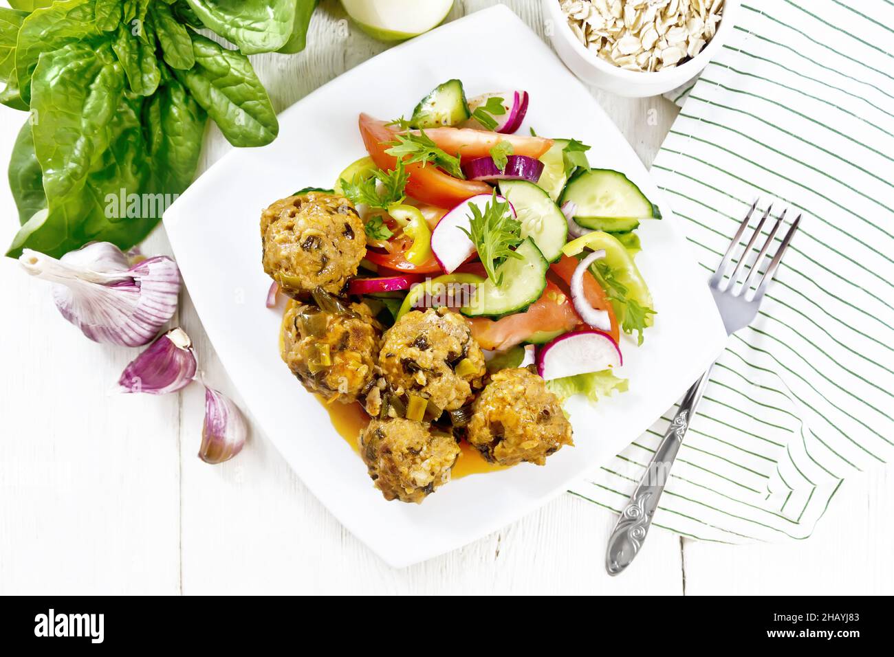 Minced meatballs with spinach, oatmeal and green onions in tomato sauce, vegetable salad in a plate, napkin on wooden board background from above Stock Photo