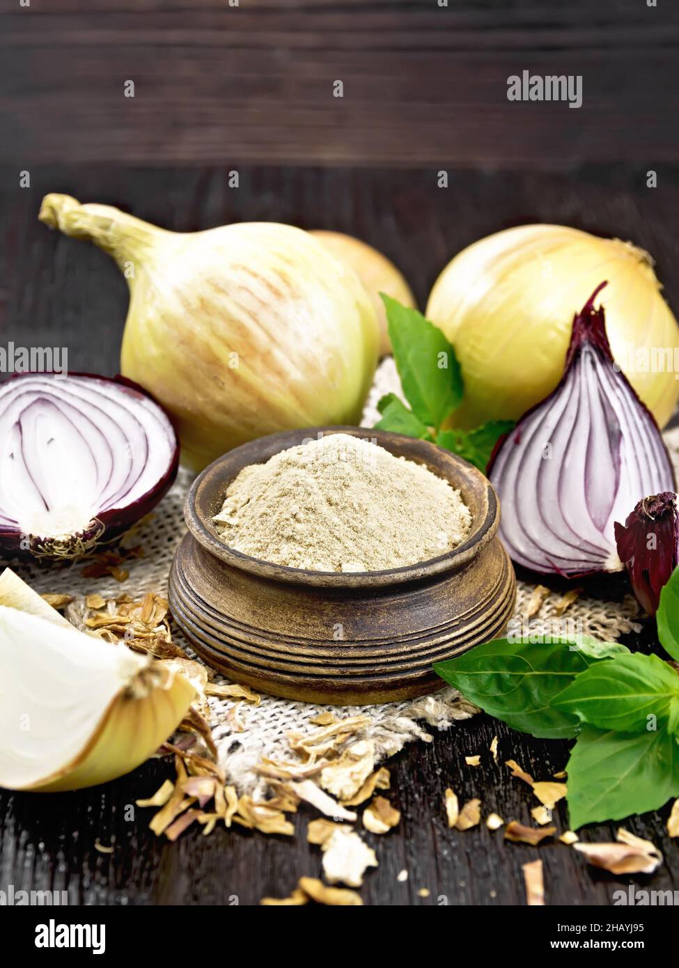 Onion powder in a bowl on a burlap napkin, purple and yellow onions, dried onion flakes and fresh basil on dark wooden board background Stock Photo