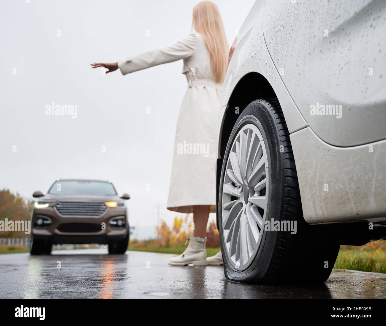 Back view of girl standing behind white vehicle with rear flat tire parked on edge of roadway and trying to stop oncoming SUV. Female driver of auto with damage tire giving stop sign to oncoming car. Stock Photo