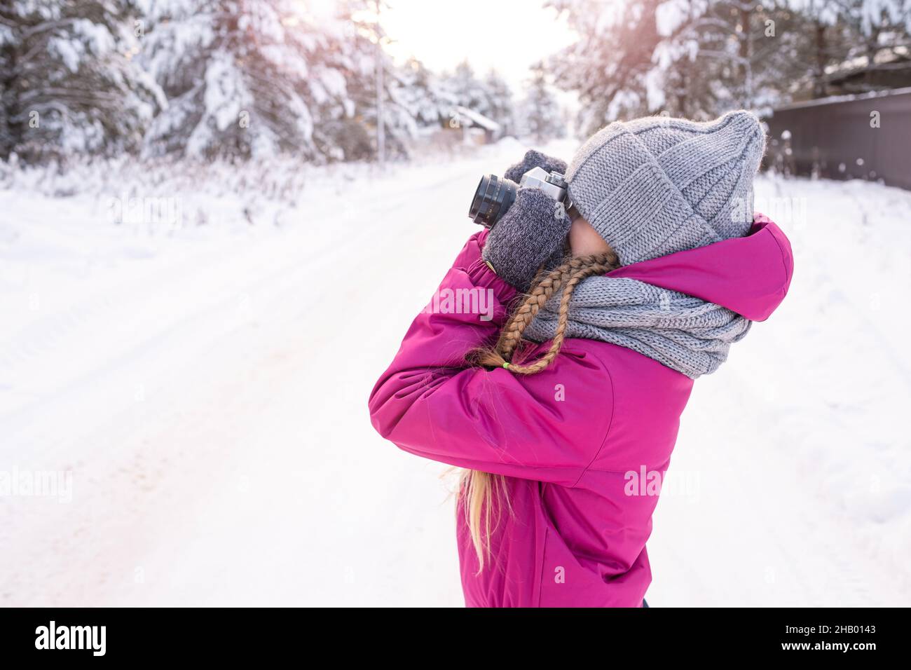 Girl in crimson winter clothes and gray knitted set is taking photo, standing sideways on winter road against background of snowy pine trees. Child Stock Photo
