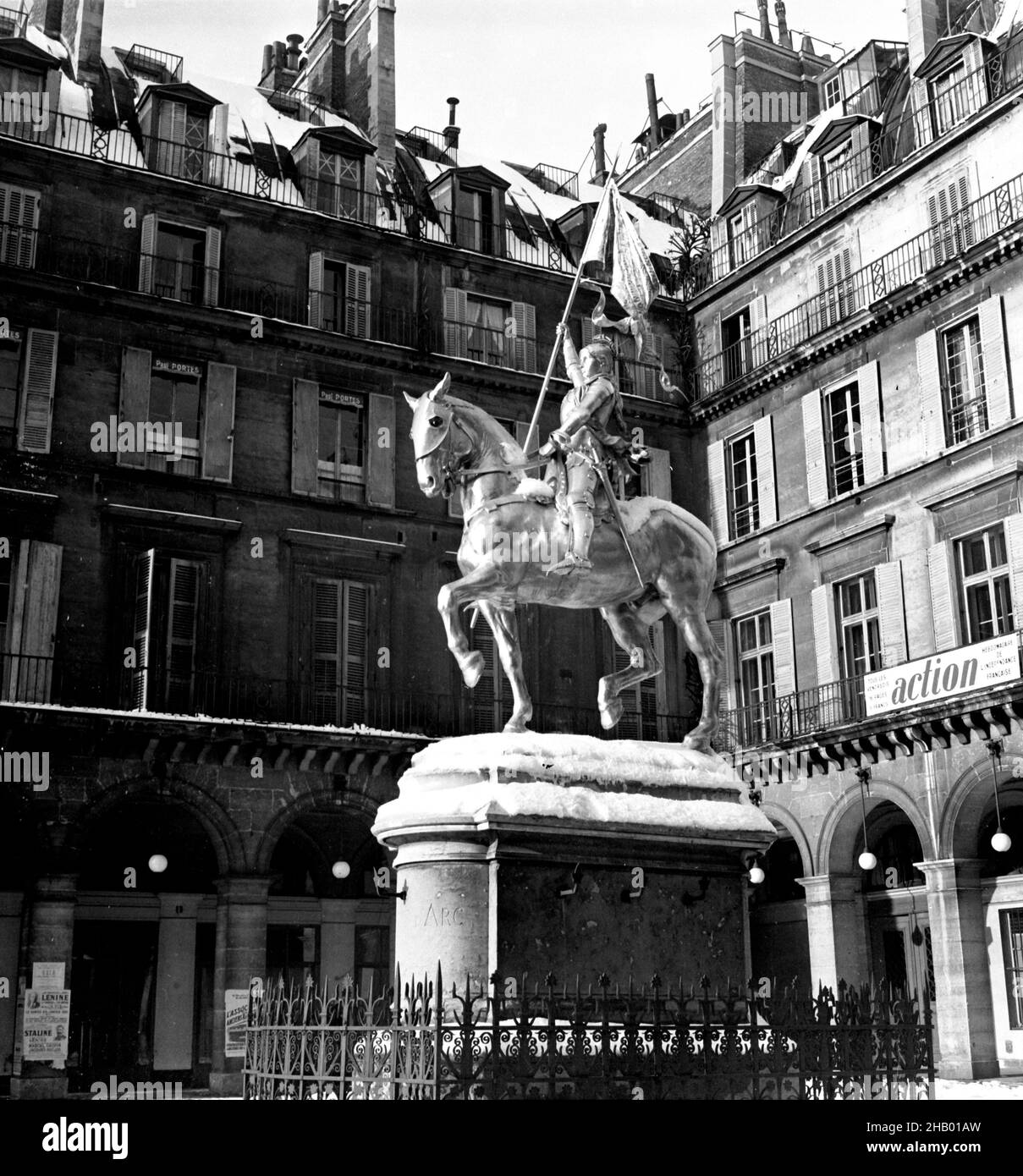 Paris Joan of Arc statue in snow in the Place des Pyramides, 1945. A photo of the bronze statue made by sculptor Emmanuel Fremiet and erected in 1874. Snow has accumulated on the back of the horse and atop the tall base that the statue sits on. There are no cars or people in this image and at first glance it might just look like a contemporary photo done in black and white. But a closer study reveals old shutters around more of the windows in the square as well as some period advertising and posted bills. A banner promotes a weekly journal called “Action”. Stock Photo