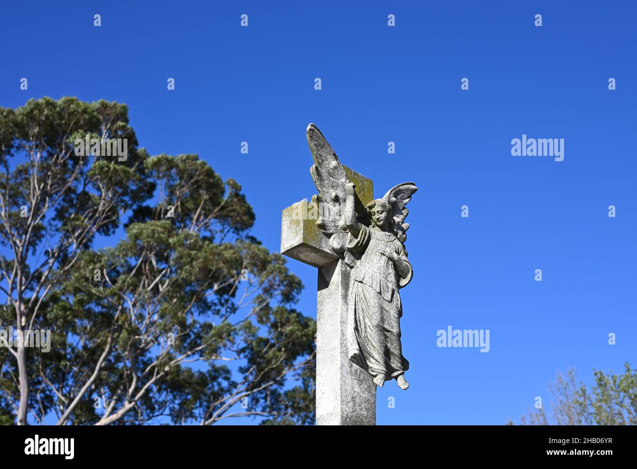 Stone sculpture of an angel in front of a cross, with the celestial being pointing towards the bright blue sky Stock Photo