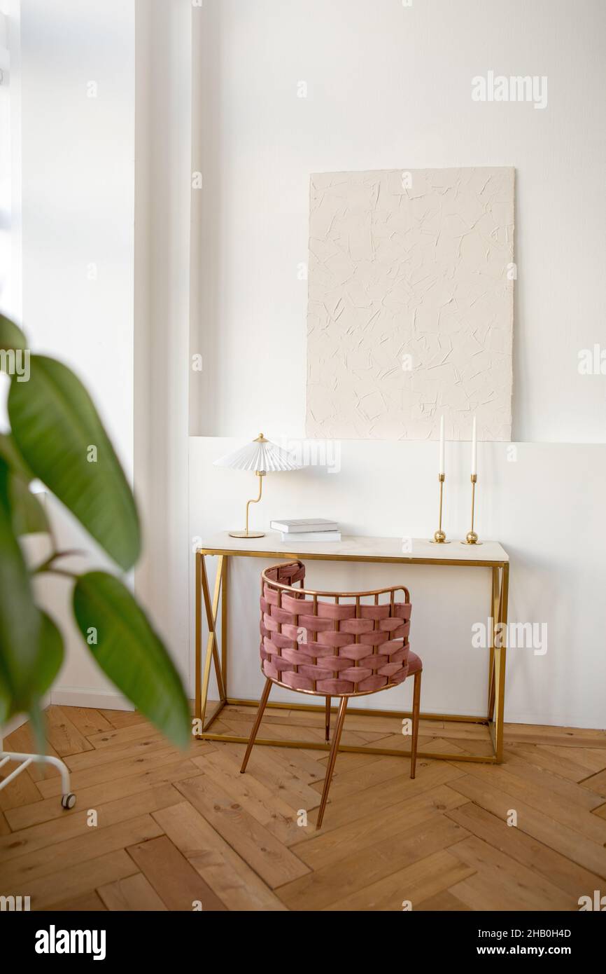 Interior design of light room furnished with table with wicker chair and lamp and books placed near white wall with ledge with white painting on it Stock Photo
