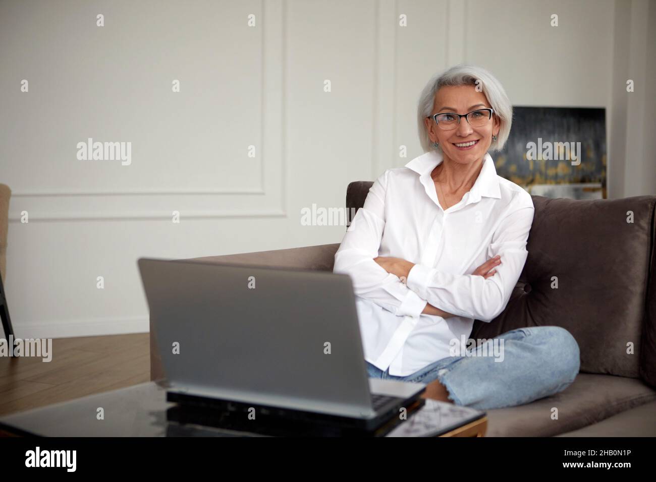 Positive female with short hair in formal clothes sitting with crossed arms on soft armchair and looking at camera while browsing laptop in light room Stock Photo