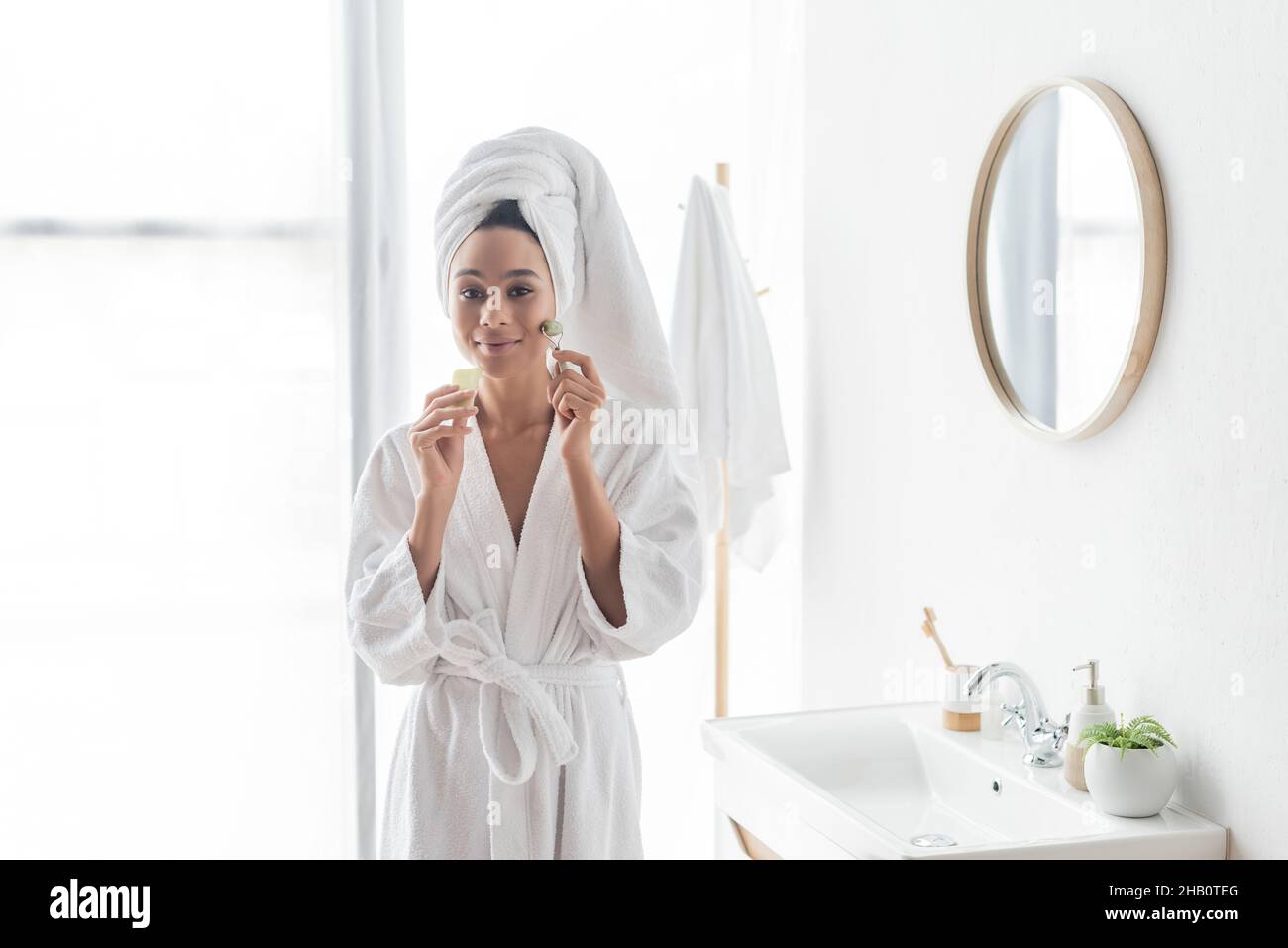 happy african american woman in bathrobe massaging face with jade ...