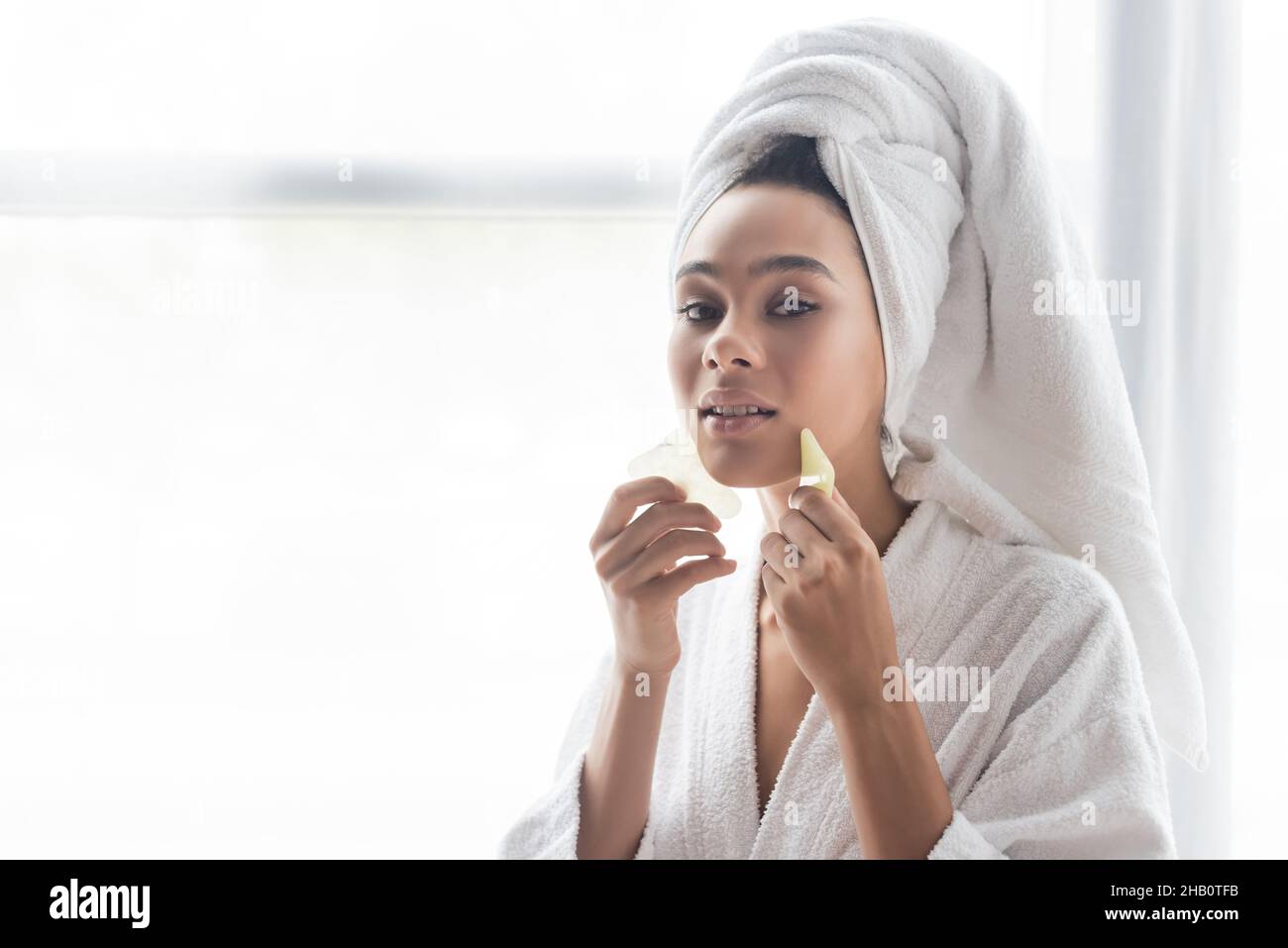 young african american woman in white towel massaging face with jade ...
