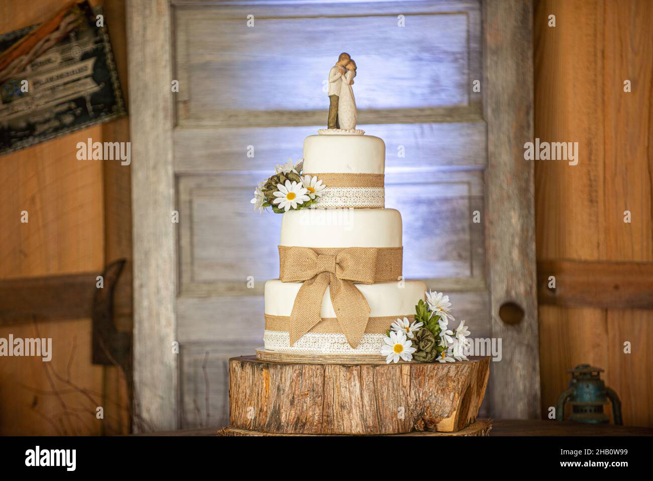 Wonderful three tier wedding cake with canvass lace and bows and sunflowers on wood bark stand and wood door background Stock Photo