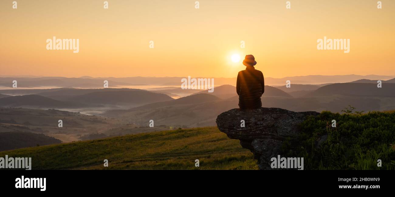 Alone tourist on the edge of the mountain hill against the backdrop of an incredible sunset mountains landscape. Panorama Stock Photo