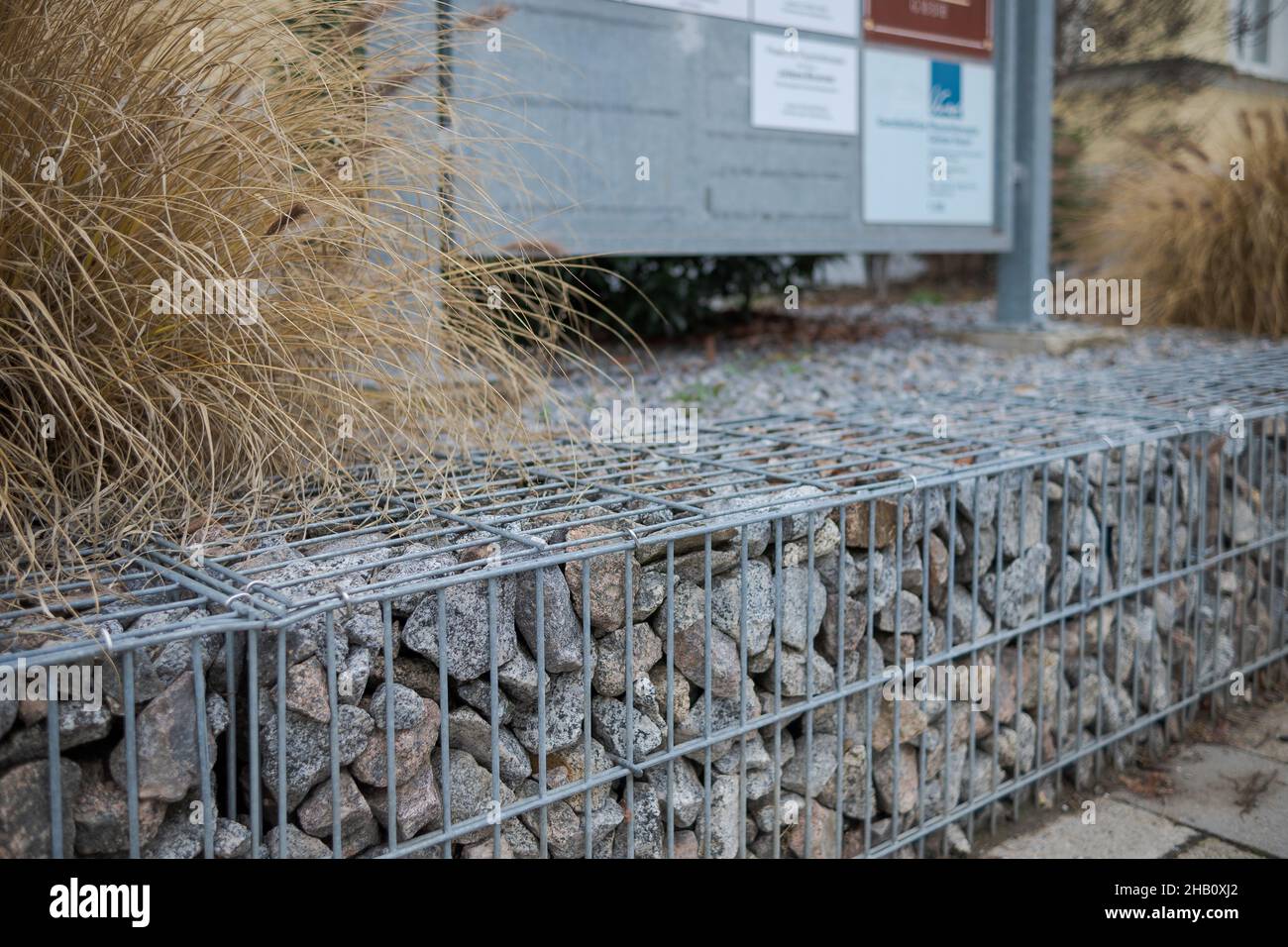 Low stylish gabion cage wall as barrier between street and house with reed grass and display sign in blurred background Stock Photo